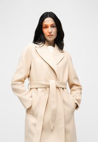 Γυναίκα με σκούρα μαλλιά που φοράει μπεζ μάλλινο παλτό με ζώνη, με τα χέρια στις τσέπες, μπροστά σε απλό φωτεινό φόντο.