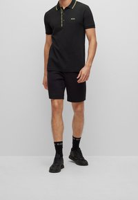 Hombre vistiendo una camiseta polo negra con ribetes amarillos, pantalones cortos negros, calcetas negras con marcas blancas y zapatillas negras, de pie contra un fondo gris.