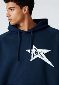 Sweat à capuche bleu marine en coton, avec un grand motif étoile blanche sur la poitrine, doté de cordons de serrage et de poignets côtelés.