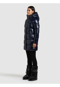 Tumši zila pūkaina virsjaka ar spīdīgu virsmu, kvadrātveida rakstu, kapuci un divām sānu kabatām, kopā ar melniem izolētiem zābakiem.