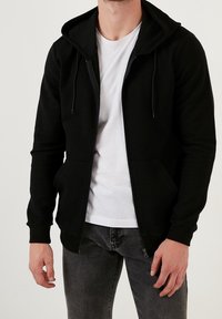 Felpa nera con zip, texture a coste, cappuccio regolabile con lacci e due tasche frontali. Indossata sopra una maglietta bianca e jeans grigi.