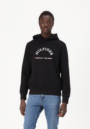 Svart hettegenser med hvit tekst "HILFIGER EST. 1985". Har ribbestrikkede mansjetter og kant, laget av mykt stoff, iført med blåjeans.