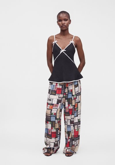 Femme portant un haut sans manches noir avec bordure blanche et un pantalon à jambes larges avec un imprimé graphique multicolore représentant divers logos de marques.
