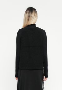 Zwarte fleece vest met een gestructureerd oppervlak, voorzien van een ronde zoom en verstelbare touwtjes aan de zijkanten. Draagt over een zwarte langemouwen shirt.