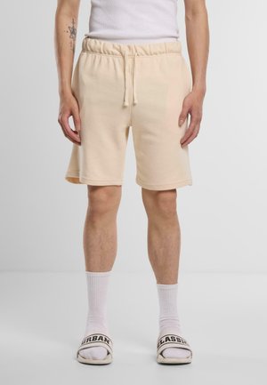 Persoon draagt beige korte broek met trekkoord, witte geribbelde sokken, witte slippers met zwarte tekst "URBAN" en "CLASSIC", en een wit hemdje.