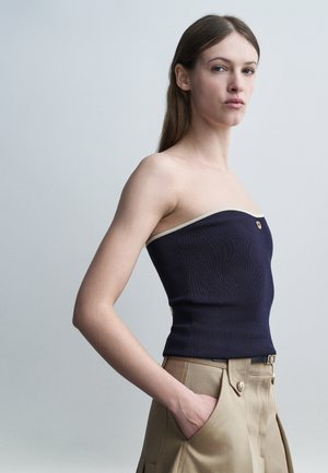 Γυναίκα με μακριά καστανά μαλλιά φοράει μπλε άνω ένδυμα χωρίς τιράντες και μπεζ παντελόνι με ψηλή μέση, στέκεται με το ένα χέρι στην τσέπη μπροστά σε γκρι φόντο.