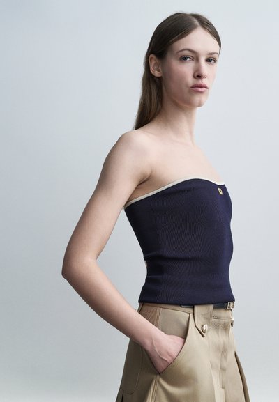 Femme aux longs cheveux bruns portant un haut bleu marine sans bretelles et un pantalon beige taille haute, debout avec une main dans la poche contre un fond gris.