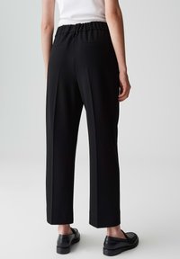 Persona vestita con pantaloni neri a vita alta e a gamba larga con tasche posteriori, top senza maniche bianco e mocassini neri, in piedi contro uno sfondo semplice.