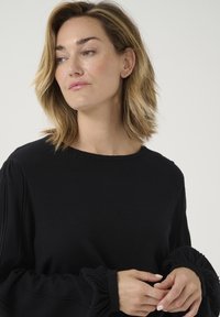Crni pulover dugih rukava s elastičnim naboranim manžetama, meke teksture, okruglim izrezom i ležernim krojem. Ruke modela su vidljive dok drži tkaninu.