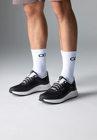 Baskets de sport noires avec semelles et lacets blancs, présentant une tige texturée et un détail de logo. Portées avec des chaussettes blanches mi-mollet affichant un logo.