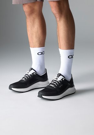 Baskets de sport noires avec semelles et lacets blancs, présentant une tige texturée et un détail de logo. Portées avec des chaussettes blanches mi-mollet affichant un logo.