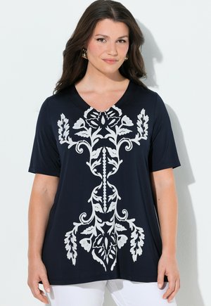 Kvinde med langt brunt hår iført guldringeøreringe og en marineblå kortærmet top med et stort hvidt blomsterprint.