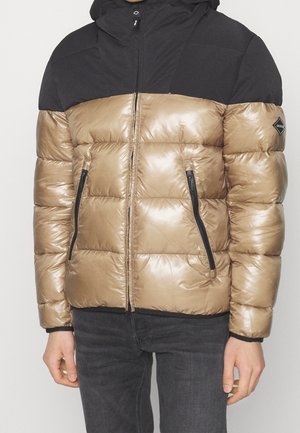 Mężczyzna w błyszczącej beżowo-czarnej kurtce puchowej z zamykanymi kieszeniami i czarnych spodniach, stojący na jednolitym tle.