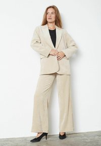 Beige kordúrová blazer a široké nohavice s jemnou textúrou. V kombinácii s čiernym topom a čiernymi podpätkami. Zobrazené na neutrálnom pozadí.
