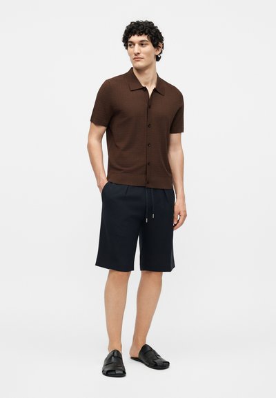 Άνδρας που φοράει καφέ κοντομάνικο πουκάμισο με κουμπιά, μπλε σκούρες βερμούδες μέχρι το γόνατο και μαύρα slip-on παπούτσια, στέκεται με τα χέρια στις τσέπες σε λευκό φόντο.