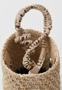 Geweven strohandtas met een ronde vorm, natuurlijke beige kleur en een getextureerd oppervlak. De handgreep is voorzien van ingewikkelde gevlochten details en accenten.