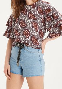 Chemise à manches courtes avec un motif bordeaux et blanc, associée à un short en denim bleu clair avec un ourlet effiloché et une ceinture noire cloutée.