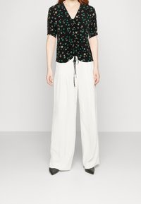 Donna che indossa una blusa nera a fiori con maniche corte arricciata abbinata a pantaloni bianchi a vita alta a gamba larga e scarpe nere a punta.