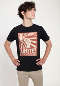 Sort bomulds t-shirt med et rødt og hvidt grafisk design, der inkluderer tekst og et hævet næve-motiv, der refererer til introversions temaer.