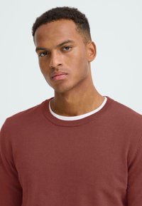 Bordó kötött pulóver bordás, kerek nyakkal, fehér pólóra rétegezve. Sima textura, hosszú ujjú és testhezálló fazon.