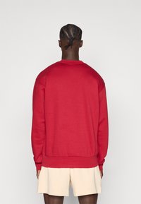 Piros pulóver bordázott mandzsettákkal és szegéllyel, kerek nyakkivágással és lazább szabással. Bézs rövidnadrággal viselve, hátulnézetből.