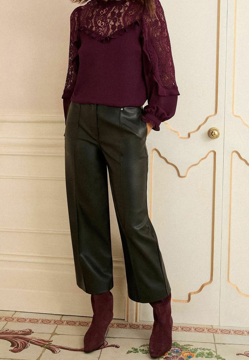 Femme debout les mains dans les poches, portant un chemisier en dentelle bordeaux, un pantalon en cuir noir coupé court et des bottines bordeaux.