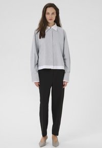 Femeie în picioare purtând un tricou lung cu mânecă de culoare gri deschis, cu guler alb, pantaloni negri și pantofi cu vârf ascuțit de culoare gri, pe un fundal simplu.