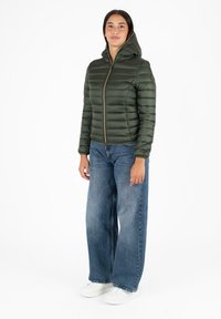 Pufferjack in donkergroen, met een capuchon, een rits aan de voorkant en een gewatteerde textuur. Gecombineerd met wijde blauwe jeans en witte sneakers.