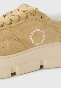 Beige semsket sko med en kraftig kremfarget såle, med flate lisser, sirkulær merkevare "COPENHAGEN STUDIOS," og teksturerte detaljer.