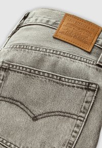 Gros plan sur la poche arrière et l’étiquette en cuir marron Levi Strauss & Co. sur un jean en denim gris clair, montrant les coutures et la texture.