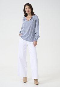 Blouse à rayures bleues et blanches à manches longues avec un col en V et des poignets froncés, associée à un pantalon large blanc et des bottes à talons beiges.