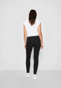 Leggings noirs au design ajusté et à la couture texturée, associés à un haut sans manches blanc. Le modèle est de dos, portant des baskets blanches.
