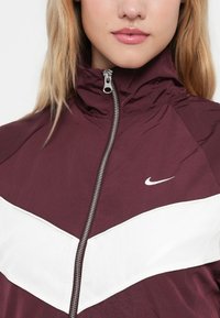 Burgundi värvi tuulekindel jakk kõrge kraega, valge chevron-riba ja hõbedane tõmblukk. Ees on väike valge Nike logo rinnal.