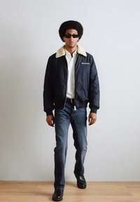 Chaqueta bomber negra con cuello de borrego beige, camisa blanca, jeans azules y zapatos negros. El modelo lleva gafas de sol oscuras y tiene un peinado afro.