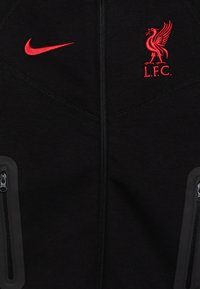 Zwarte ritsjack van zacht materiaal. Voorzien van een rood Nike swoosh en het Liverpool FC-logo. Twee zijzakken met rits en textuurdetails.