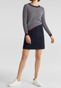 Granatowa bluzka w białe groszki z długim rękawem i okrągłym dekoltem; zestawiona z ciemnogranatową spódnicą w kształcie litery A oraz białymi trampkami.