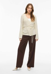 Krēmkrāsas wrap stila kardigans no mīksta materiāla ar garām piedurknēm un sasiešanas aizdari. Kombinēts ar tumši brūnām plaša silueta biksēm un bēšiem augstpapēžu apaviem.