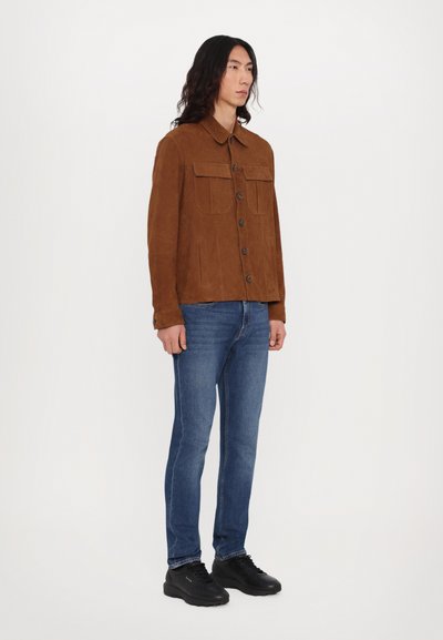 Brun ruskindsjakke med krave, to brystlommer og knaplukning, parret med blå denim jeans og sorte sneakers.