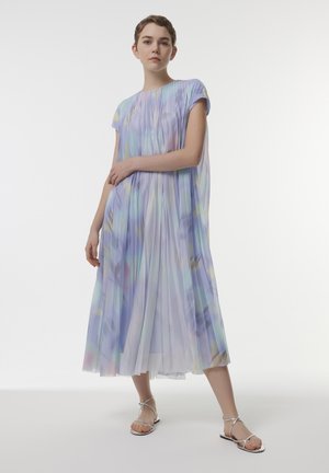 Lichtgewicht, geplooid jurkje in pastelkleuren van lila, blauw en geel. Korte mouwen, knielang met een vloeiende silhouette, gecombineerd met zilveren sandalen.