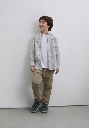 Camicia a quadri chiara con bottoni sopra una t-shirt bianca, abbinata a pantaloni beige e sneakers verdi con chiusura a strappo. Design semplice e colori neutri.