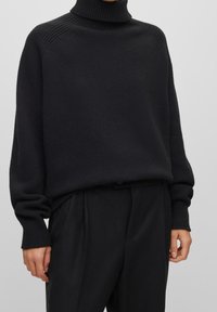 Svart stickad polotröja med ribbdetaljer vid halsen och axelsömmarna, kombinerad med skräddarsydda svarta byxor.