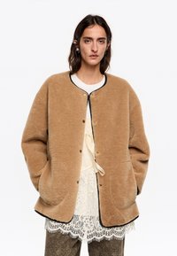 Mujer que lleva una chaqueta de felpa marrón sobre una blusa de encaje blanca y pantalones oscuros, con las manos en los bolsillos de la chaqueta, expresión neutral y un clip en el cabello en el lado izquierdo.