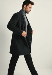 Mężczyzna w ciemnym płaszczu i szarym golfie, idący z rękami poprawiającymi płaszcz na jednolitym jasnym tle.
