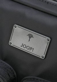 Borsa in tessuto nero con un'etichetta metallica rettangolare con il nome del marchio "JOOP!" e un emblema grafico sopra, fissata con quattro rivetti.