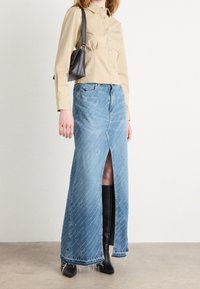 Kvinna som bär en beige knappad skjorta, lång blå denimkjol med framfickor, svarta knähöga stövlar och en svart axelväska mot en vit vägg.