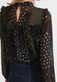Schwarze, transparente Bluse mit floralem, herzförmigem und gepunktetem Muster. Verfügt über einen Rüschenausschnitt und einen Schlüssellochausschnitt mit Knopfverschluss.