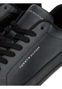 Gros plan d'une basket en cuir noir avec des lacets noirs et le logo blanc Tommy Hilfiger ainsi que le drapeau sur le côté et la languette.