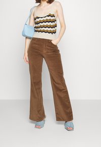 Hnědé manšestrové kalhoty s rozšířenými nohavicemi spojené s vzorovaným tílkem s vlnitými pruhy v modré, žluté a krémové barvě. Světle modrá taška doplňuje vzhled.