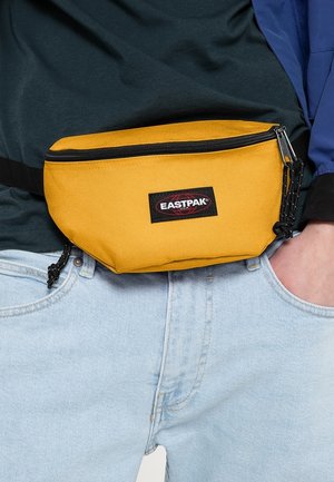 Marsupio in tessuto giallo con cerniera nera, toppa con marchio sulla parte anteriore e tracolla regolabile, indossato in vita sopra jeans azzurri.