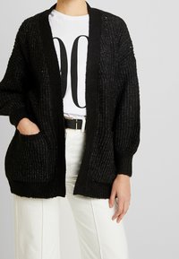 Svart stickad cardigan med öppen framdel, långa ärmar, ribbad textur och två framfickor. Bärs över en vit t-shirt och ljusa byxor.